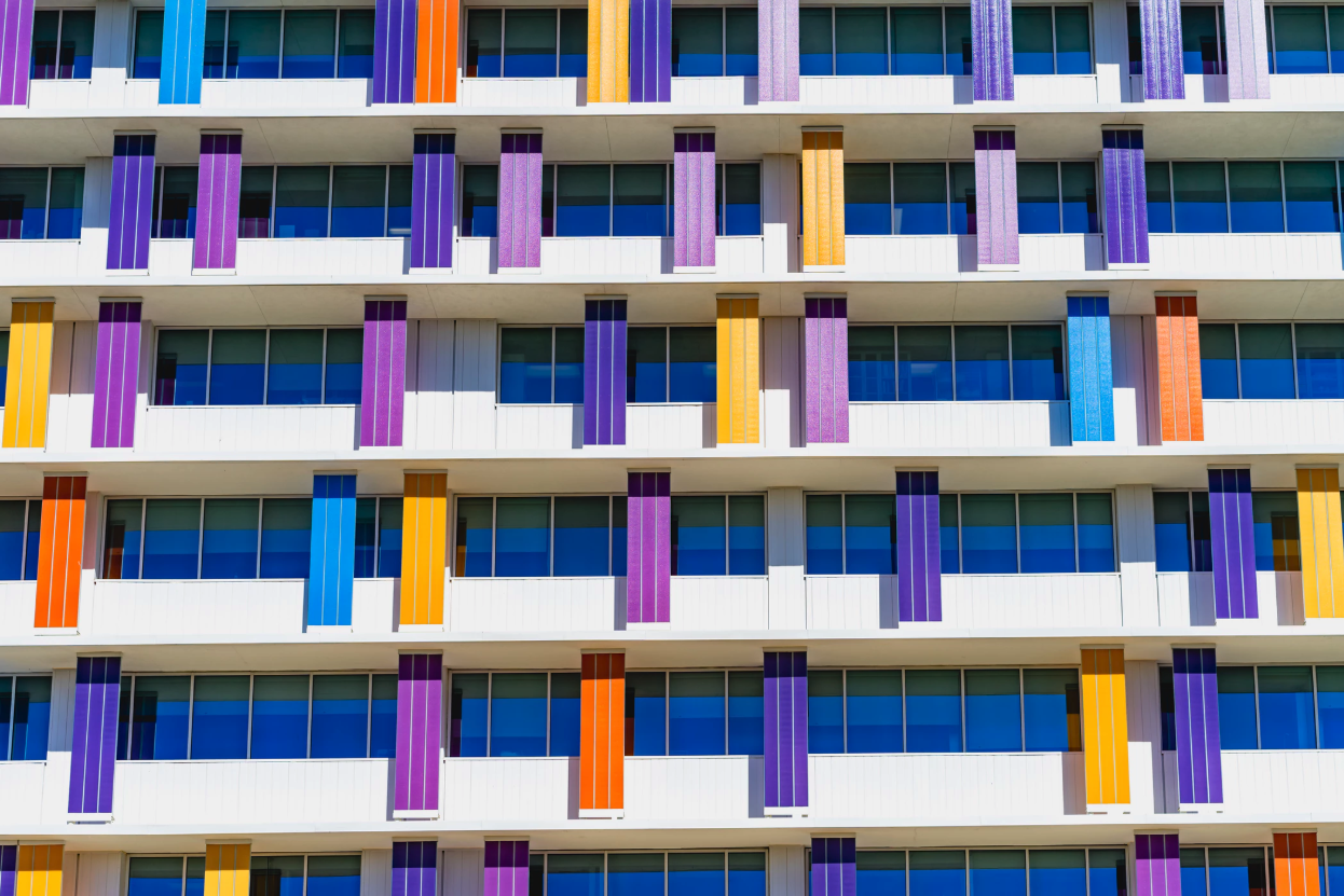 The side of a multistory building with lots of windows and the support pillars are teal, peach, purple, magenta and orange.
